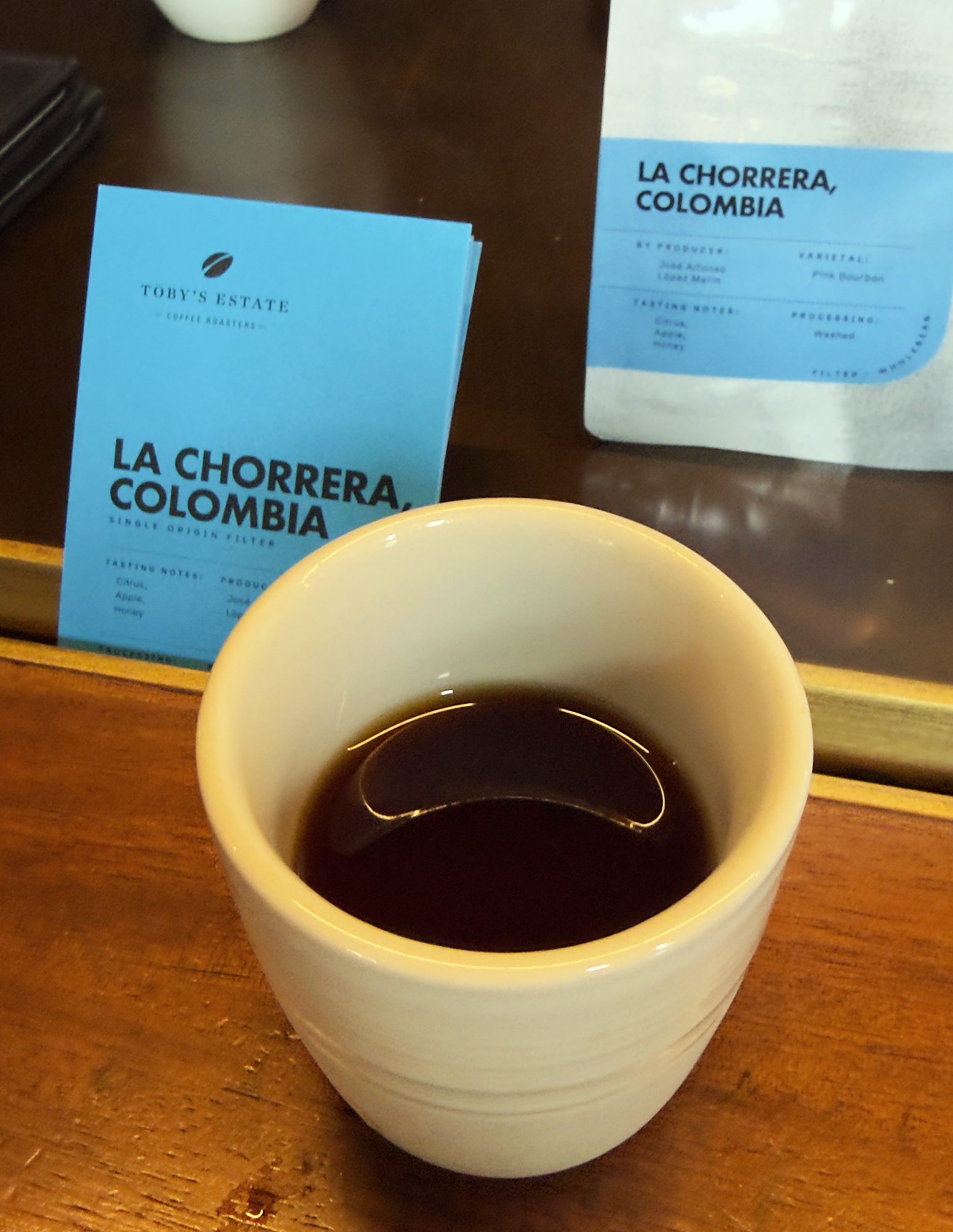 A cup of filter coffee on a counter with its lot card and bag behind, both reading La Chorrera, Colombia, Pink Bourbon, washed, citrus, apple, honey